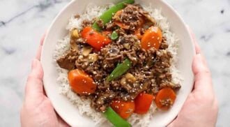 A bowl of white rice topped with a savory ground beef stir fry, featuring carrots, snap peas, mushrooms, and sesame seeds, held in two hands.