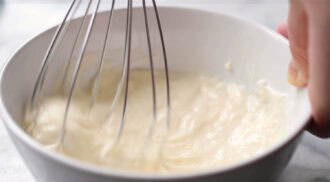 A hand uses a wire whisk to mix a thick, creamy batter in a white bowl, perfect for coating honey walnut shrimp.