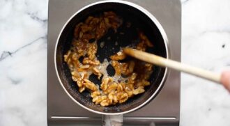 A person stirs walnuts coated in caramelized sugar in a small saucepan on a stovetop, preparing the sweet, crunchy nuts often used as a topping for honey walnut shrimp.