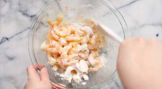 A person prepares honey walnut shrimp by mixing raw shrimp with a white powder, likely cornstarch or flour, in a clear glass bowl on a marble countertop.