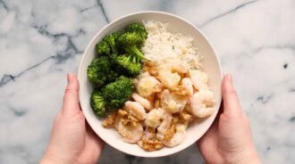 A bowl of honey walnut shrimp with garlic sauce, steamed broccoli, and white rice, held by two hands on a marble surface.