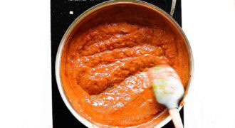 A saucepan filled with thick red chicken tikka masala sauce is being stirred with a spatula on a black stovetop.