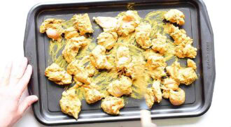 Raw chicken pieces coated in a yellow chicken tikka masala marinade are spread out on a black baking tray, with a hand using a spatula to arrange them.