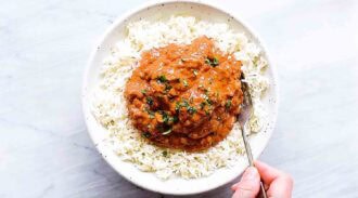 A bowl of white rice topped with a serving of orange-brown chicken tikka masala, garnished with herbs; a hand holds a fork in the bowl.