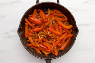 Sliced red bell peppers and onions sautéed in a dark skillet on a white marble surface, perfect as a flavorful base for shrimp fajitas.