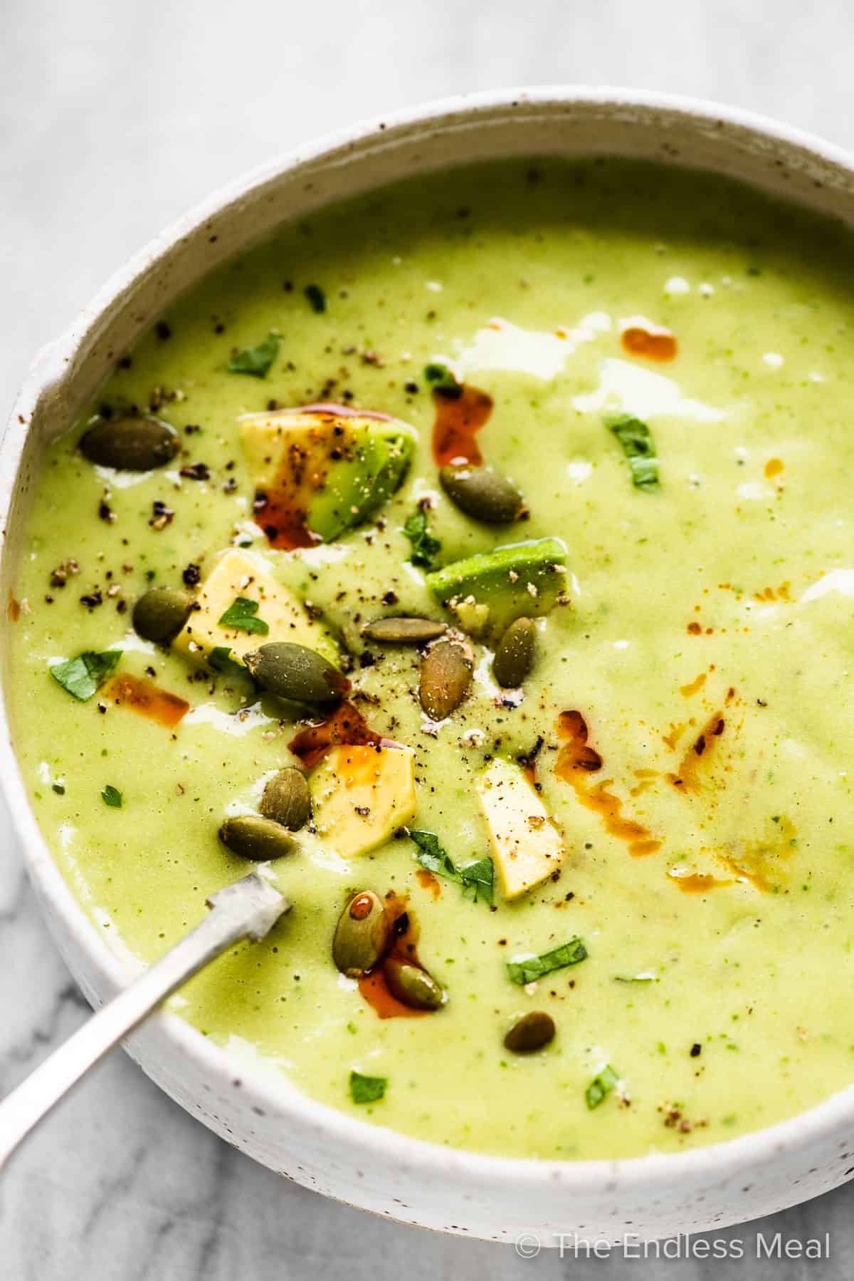A spoon in a bowl of avocado soup topped with diced avocados and pumpkin seeds.