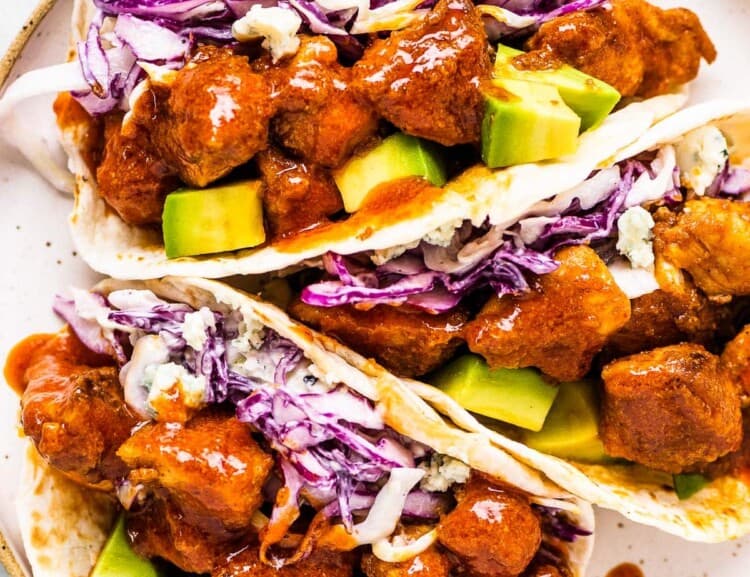 Three buffalo chicken tacos on a plate with coleslaw and avocados.