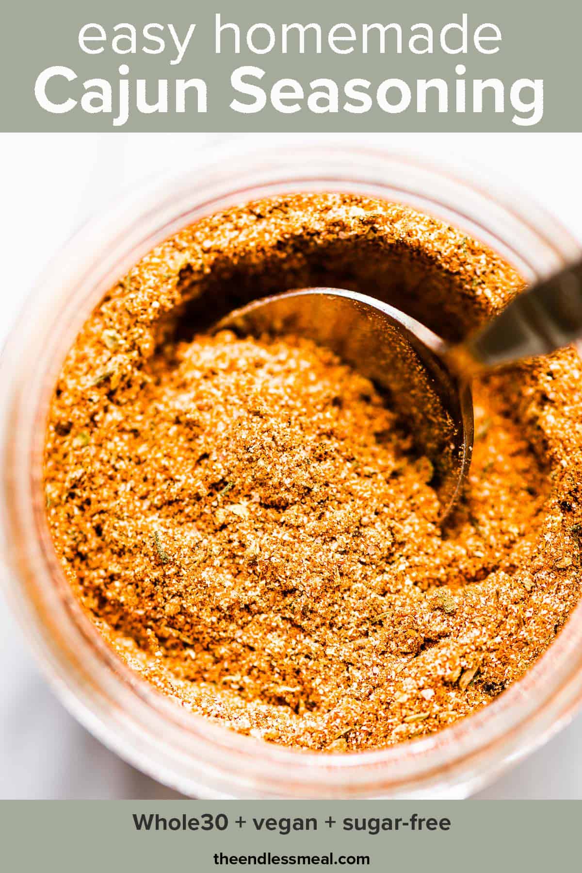 Homemade cajun seasoning in a glass jar with a spoon with the recipe title on top of the picture.