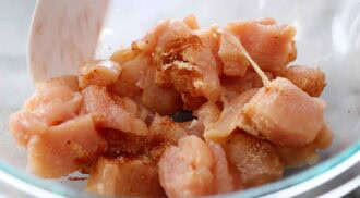 Raw, cubed chicken pieces in a glass bowl, sprinkled with spices for buffalo chicken tacos, being stirred with a white utensil.