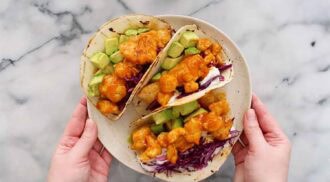 A plate with three buffalo chicken tacos filled with cauliflower, avocado chunks, and shredded purple cabbage, held by two hands over a marble surface.