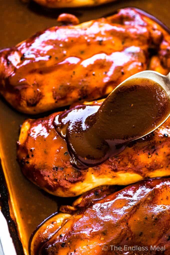 BBQ sauce being spooned over the top of baked chicken in a pan.