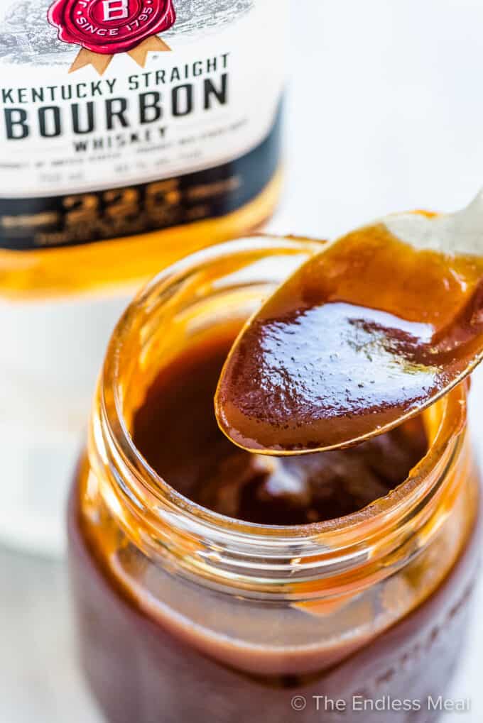 A jar of bourbon bbq sauce with a bottle of bourbon in the background.
