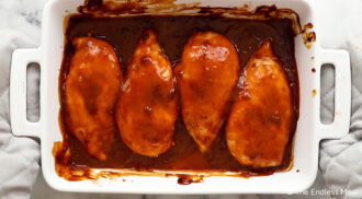 Four baked chicken breasts coated in barbecue sauce in a white ceramic baking dish.