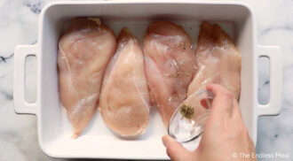 A hand sprinkles black pepper from a small glass bowl onto four raw chicken breasts in a white rectangular baking dish.