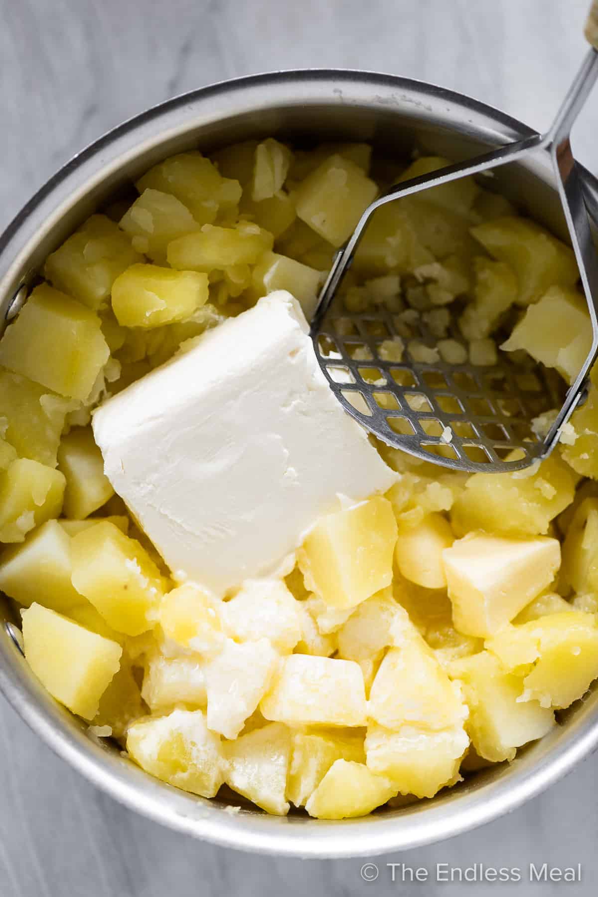 Cooked potatoes in a pot with cream cheese and a potato masher.