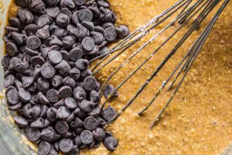 A metal whisk rests in a bowl of brown batter for pumpkin oatmeal muffins, with a pile of chocolate chips about to be mixed in.