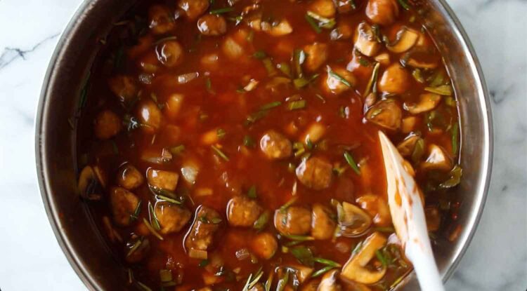 A saucepan filled with vegan mushroom gravy, tomato-based sauce, chopped mushrooms, onions, and herbs is being stirred with a white spatula on a marble countertop.