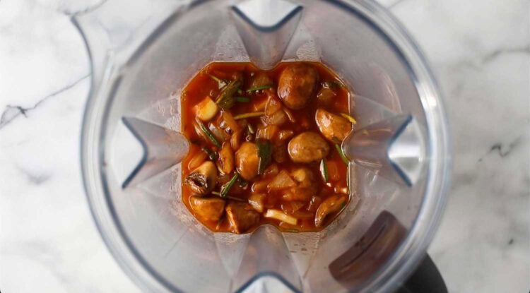 Overhead view of a blender containing whole mushrooms, chopped onions, herbs, and a reddish sauce on a marble countertop, ready to be blended into vegan mushroom gravy.