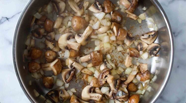 Chopped mushrooms and diced onions, perfect for vegan mushroom gravy, cooking in a stainless steel pan on a marble countertop.