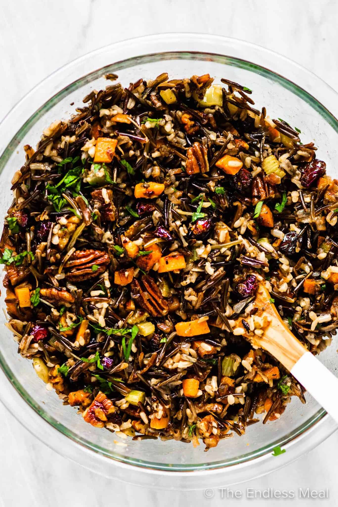 Wild rice stuffing with pecans, sweet potatoes, and cranberries in a glass bowl with a wooden spoon.