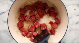 Chunks of raw beef, soon to be transformed into a flavorful Thai beef curry, are being seared in a large white pot on the stovetop, with a spatula visible stirring the meat.