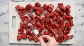 Diced raw beef cubes for Thai beef curry are spread on a white cutting board while a hand sprinkles salt over them with a measuring spoon.