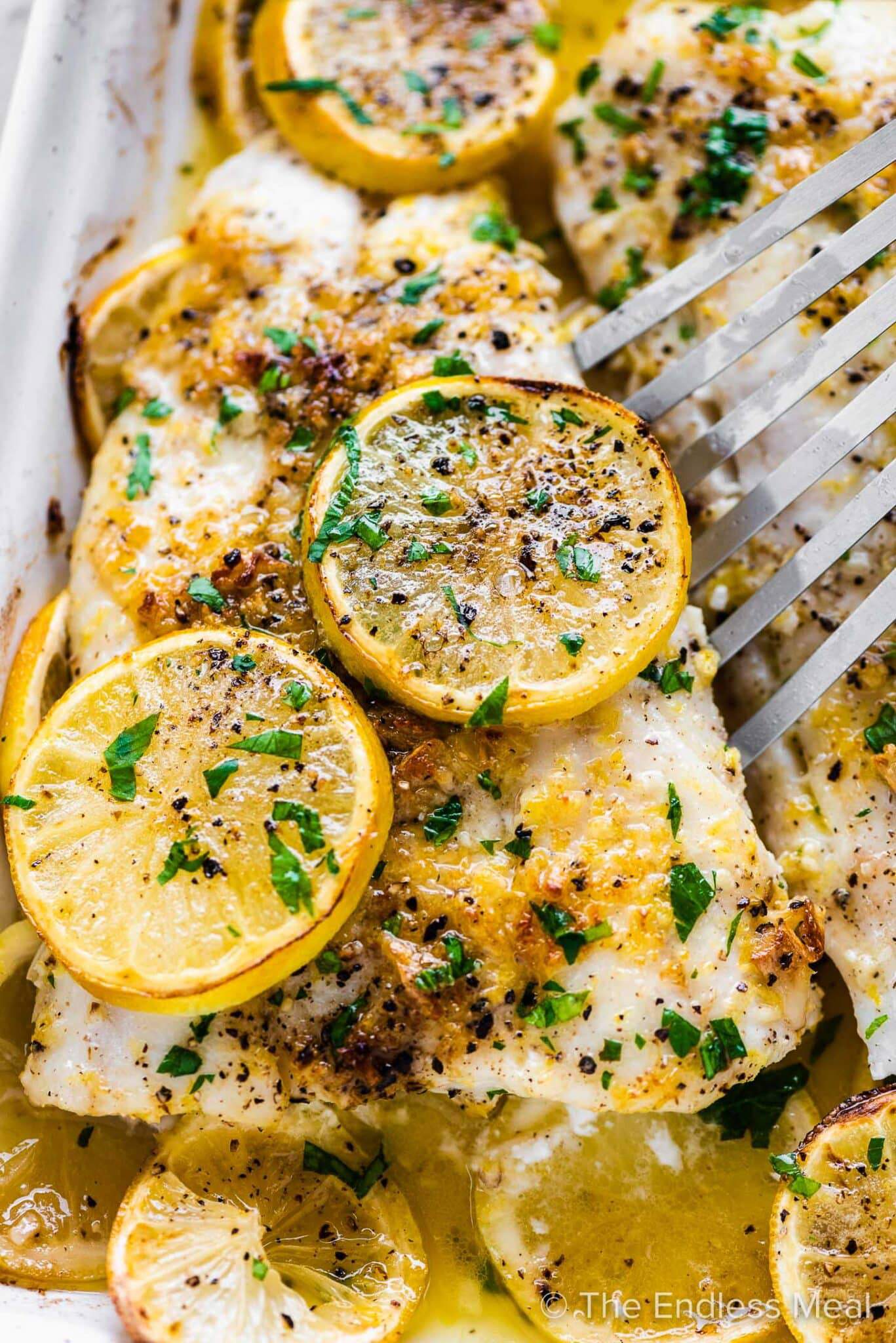 A spatula lifting a piece of baked fish with lemons out of a baking dish.