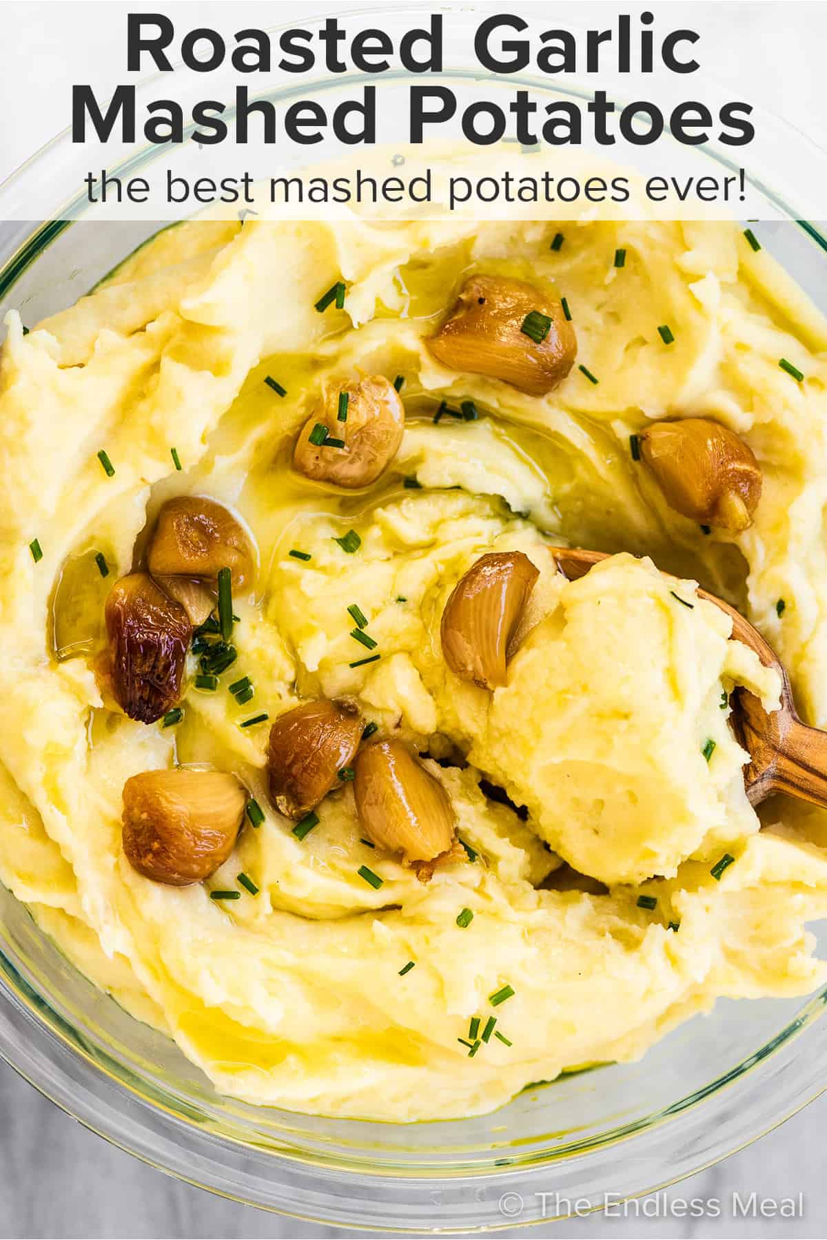 A serving bowl of roasted garlic mashed potatoes with a wooden spoon in it and the recipe title on top of the picture.