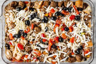 A glass baking dish filled with a breakfast strata made of bread cubes, sausage, chopped red peppers, black olives, and shredded cheese.
