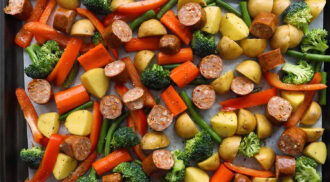 Sheet Pan Sausage and Veggies features sliced sausage, potatoes, carrots, green beans, red bell peppers, and broccoli arranged on a baking sheet, ready to be cooked.