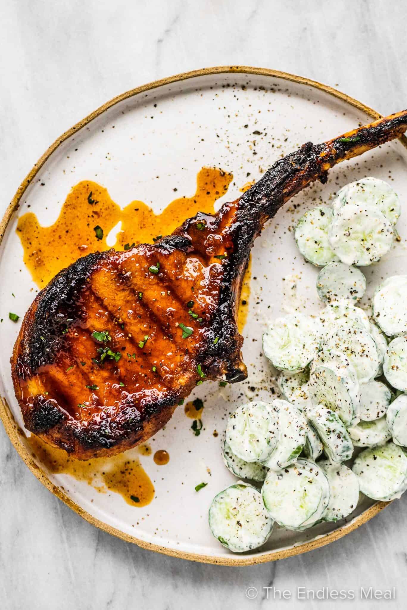 A tomahawk pork chop with honey garlic sauce on a plate with cucumber salad.
