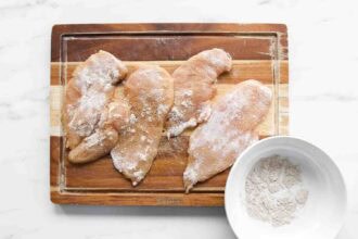 Raw chicken breasts coated in flour are arranged on a wooden cutting board, ready to be transformed into a delicious Creamy Cajun Chicken dish, with a white bowl of flour nearby.