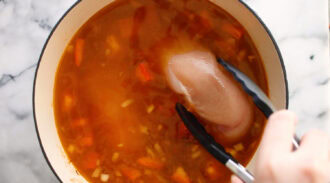 A hand uses tongs to add a raw chicken breast to a pot of simmering Chicken Taco Soup with chopped vegetables on a marble countertop.