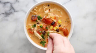 A hand sprinkles herbs onto a bowl of creamy Chicken Taco Soup containing shredded chicken, corn, black beans, tomatoes, and garnishes.