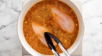 Tongs placing raw chicken breasts into a pot of seasoned broth on a stovetop.