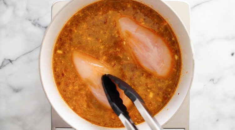 Tongs placing raw chicken breasts into a pot of seasoned broth on a stovetop.