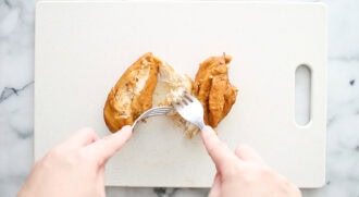 Two hands use a fork and knife to shred cooked chicken breast on a white cutting board.