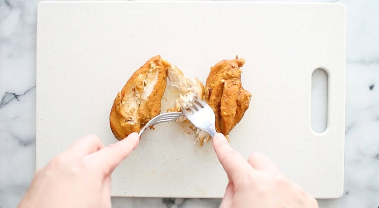 Two hands use a fork and knife to shred cooked chicken breast on a white cutting board.
