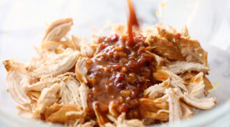 Shredded chicken in a glass bowl with a thick brown sauce being poured over it.