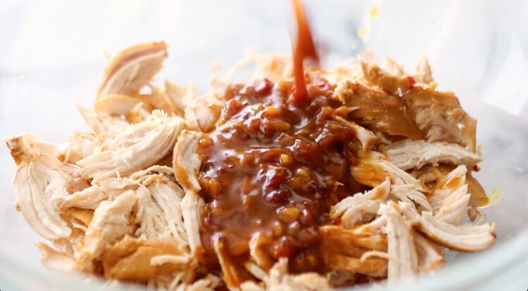 Shredded chicken in a glass bowl with a thick brown sauce being poured over it.