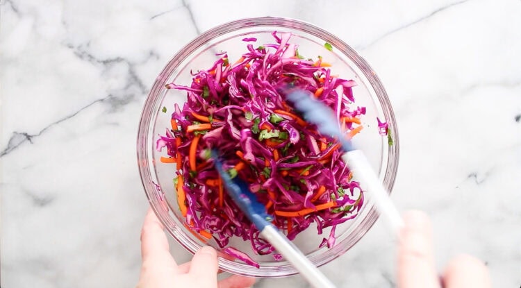 A glass bowl of shredded red cabbage, carrots, and herbs is being mixed with tongs on a marble surface.