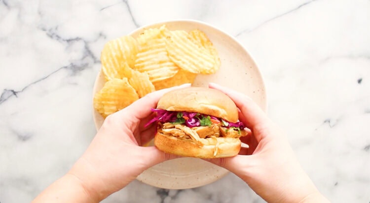 Two hands hold a sandwich with shredded meat and purple cabbage above a beige plate with ridged potato chips on a white marble surface.