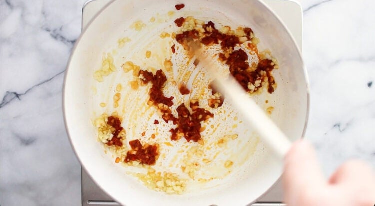 A hand stirs minced garlic and red chili paste in a white pan on a stovetop.