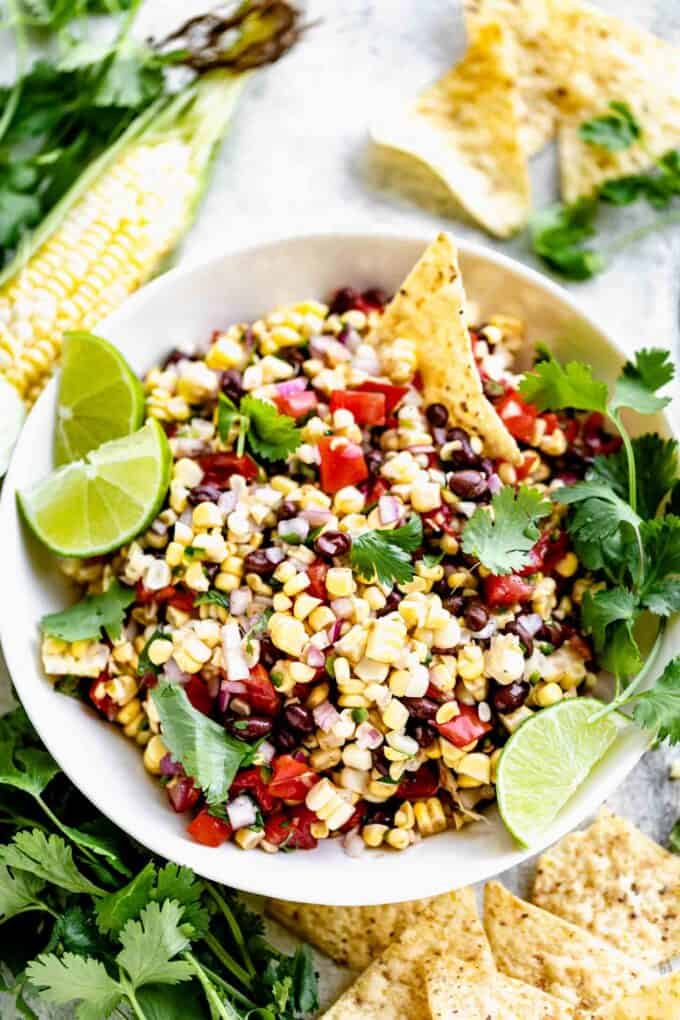a bowl of corn salsa with a chip in it, garnished with cilantro sprigs and lime wedges