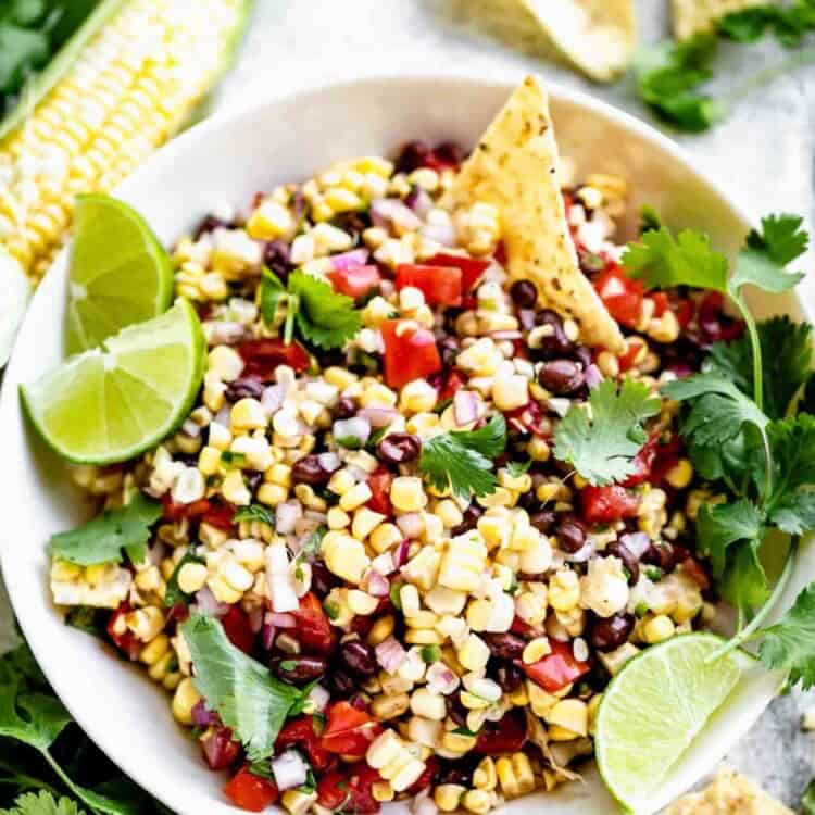 a bowl of corn salsa with a chip in it, garnished with cilantro sprigs and lime wedges