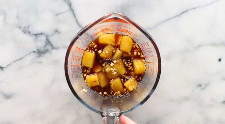 A glass measuring cup filled with pineapple chunks, minced garlic, and a dark liquid marinade—perfect for preparing juicy pineapple pork chops—sits on a marble countertop.
