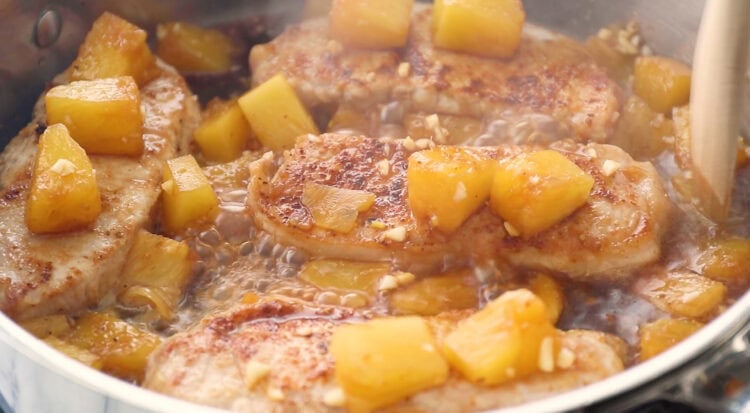 Pineapple pork chops are pan-cooked with chunks of pineapple and minced garlic in a shallow sauce, creating a golden and caramelized appearance.