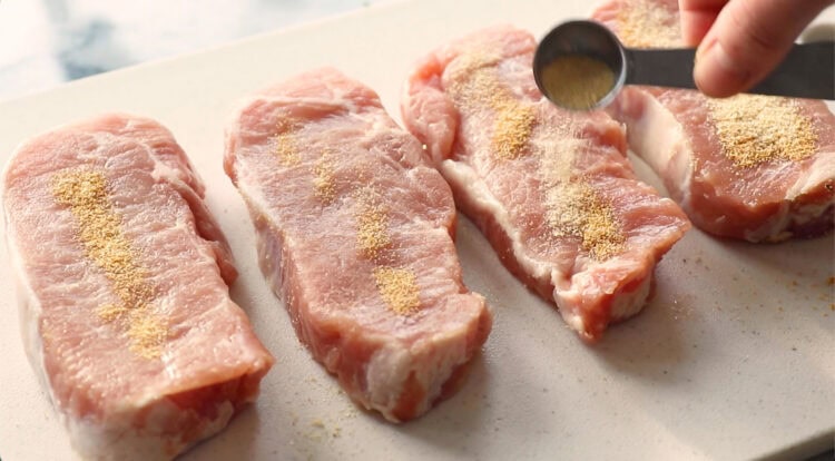 Four raw pineapple pork chops on a cutting board are being sprinkled with seasoning from a measuring spoon.