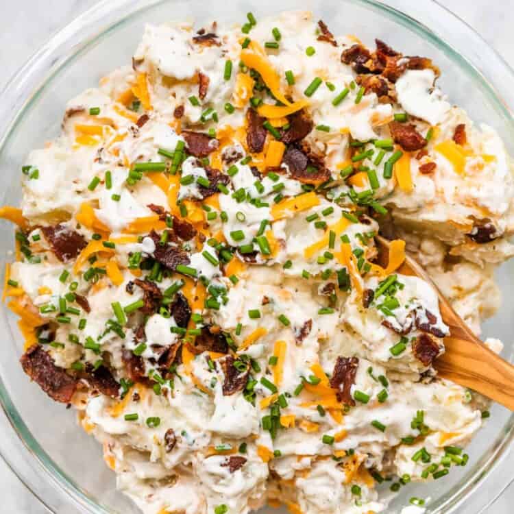 Loaded baked potato salad in a glass bowl with a wooden spoon.