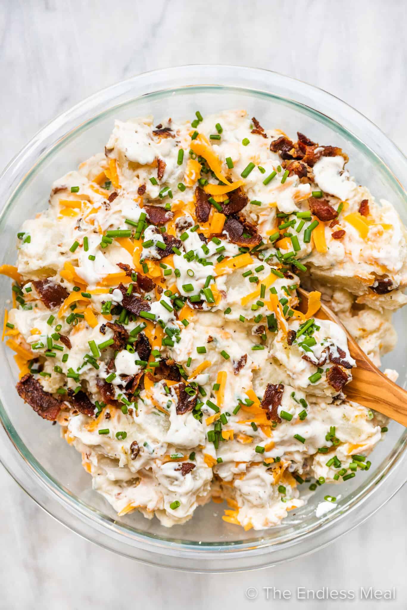 Loaded baked potato salad in a glass bowl with a wooden spoon.